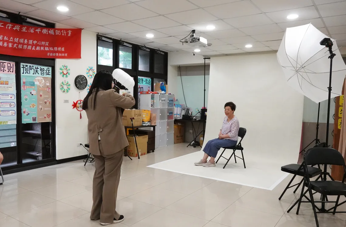 側拍：榕榕站在燈旁，替坐在布幕前的奶奶拍照